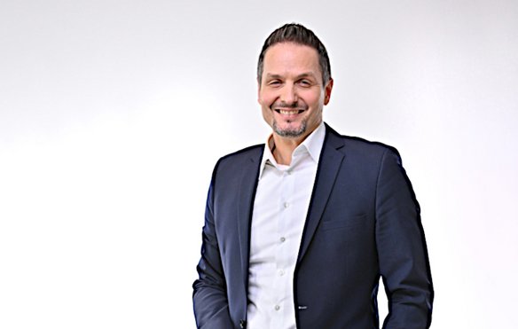 Mann in dunklem Anzug mit weißem Hemd, lächelt in die Kamera. Er hat kurze braune Haare und einen gepflegten Bart. Der Hintergrund ist neutral und hell.