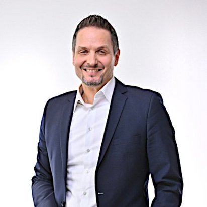 Mann in dunklem Anzug mit weißem Hemd, lächelt in die Kamera. Er hat kurze braune Haare und einen gepflegten Bart. Der Hintergrund ist neutral und hell.