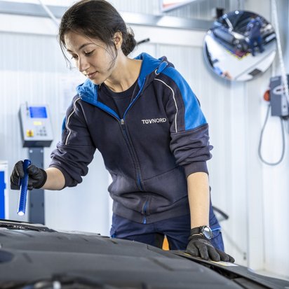 tuev-nord-fahrzeugpruefung-werkstatt Eine junge Frau mit dunklen Haaren und einer blauen TÜV NORD-Arbeitsjacke inspiziert den Motorraum eines Autos in einer Werkstatt. Sie trägt schwarze Schutzhandschuhe und hält eine Taschenlampe in der Hand, während sie sich konzentriert über den offenen Motorraum beugt. Im Hintergrund sind Werkstattausrüstung, technische Geräte und ein runder Spiegel zu sehen, der einen Teil der Werkstatt reflektiert.