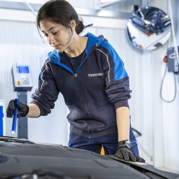 Eine junge Frau mit dunklen Haaren und einer blauen TÜV NORD-Arbeitsjacke inspiziert den Motorraum eines Autos in einer Werkstatt. Sie trägt schwarze Schutzhandschuhe und hält eine Taschenlampe in der Hand, während sie sich konzentriert über den offenen Motorraum beugt. Im Hintergrund sind Werkstattausrüstung, technische Geräte und ein runder Spiegel zu sehen, der einen Teil der Werkstatt reflektiert.