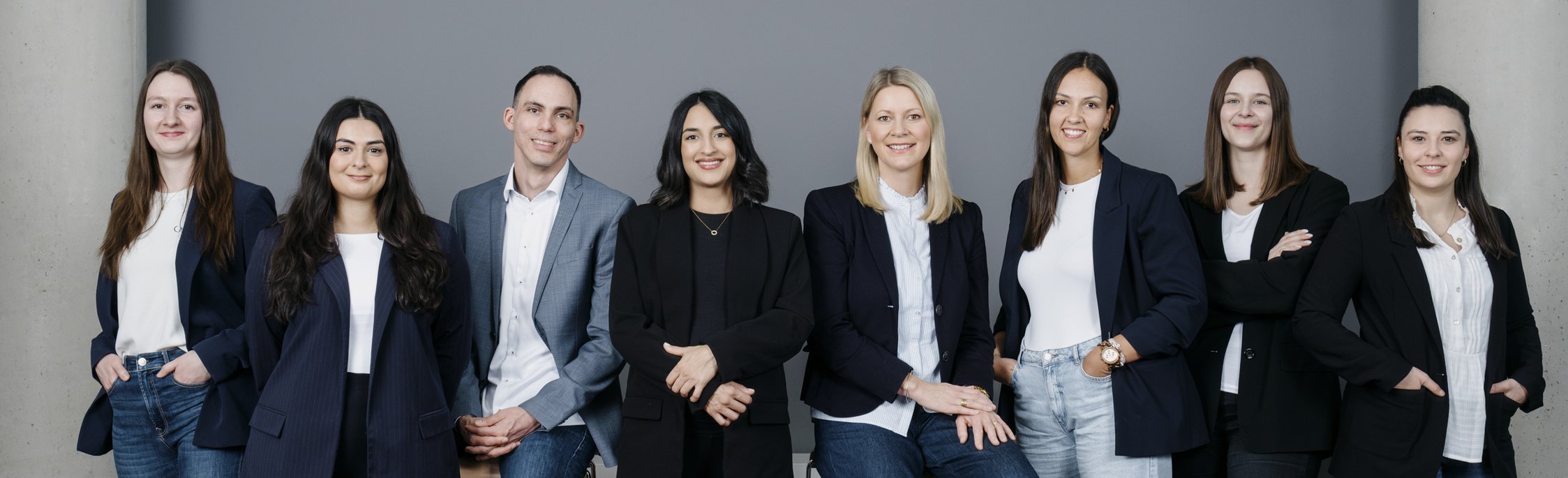 Teamfoto vor neutralem Hintergrund Acht Personen in Business-Kleidung stehen nebeneinander vor einem grauen Hintergrund und lächeln.