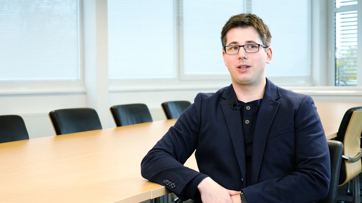 Das Bild zeigt Brian Niehoefer, Evaluator mit Schwerpunkt auf IT-Sicherheitsprodukte, in einem hellen Konferenzraum. Er trägt ein dunkelblaues Sakko über einem schwarzen Shirt mit Knopfleiste. Seine Brille und der entspannte, leicht seitliche Blick vermitteln Offenheit und Fachkompetenz. Er sitzt an einem großen Konferenztisch mit schwarzen Stühlen. Die Fenster im Hintergrund sind mit Jalousien abgedeckt, die diffuses Tageslicht in den Raum lassen. Das Bild gehört zu einem Mitarbeiter-Testimonial und vermittelt eine professionelle, sachliche Atmosphäre.