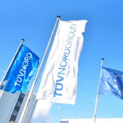 Drei Fahnen wehen vor einem blauen Himmel: eine blaue TÜV NORD-Fahne mit weißer Schrift, eine weiße TÜV NORD GROUP-Fahne mit blauer Schrift und eine blaue EU-Fahne mit gelben Sternen. Im Hintergrund sind moderne Bürogebäude zu sehen.