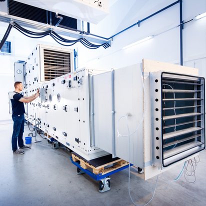 Ein Techniker in blauer Arbeitskleidung arbeitet an einer großen weißen Klimaprüfkammer in einem Labor. Die Kammer ist mit zahlreichen Messinstrumenten ausgestattet und steht auf Rollwagen. Kabel und Rohre verlaufen entlang der Decke und Wände des hell erleuchteten Raums