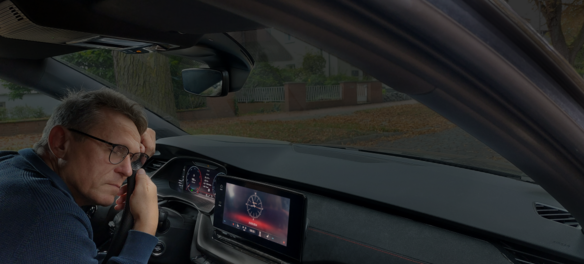 Ein Mann mit Brille und dunkler Kleidung sitzt erschöpft oder nachdenklich am Steuer eines Autos. Er lehnt seinen Kopf auf seine Hand und schaut müde nach vorne. Das Armaturenbrett zeigt eine digitale Uhr auf dem Bildschirm des Infotainmentsystems. Durch die Windschutzscheibe sind eine herbstliche Straße mit Laub sowie ein Wohngebiet mit Häusern und einem Gartenzaun zu sehen.