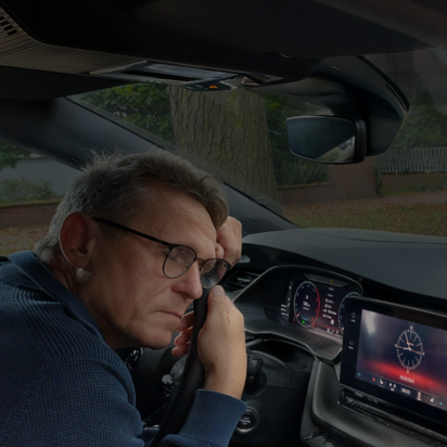 Ein Mann mit Brille und dunkler Kleidung sitzt erschöpft oder nachdenklich am Steuer eines Autos. Er lehnt seinen Kopf auf seine Hand und schaut müde nach vorne. Das Armaturenbrett zeigt eine digitale Uhr auf dem Bildschirm des Infotainmentsystems. Durch die Windschutzscheibe sind eine herbstliche Straße mit Laub sowie ein Wohngebiet mit Häusern und einem Gartenzaun zu sehen.