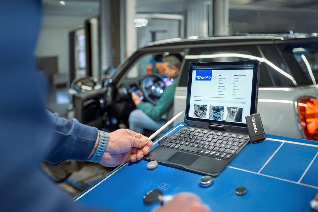 Tablet mit Fahrzeuginformationen in der Werkstatt, während ein Auto im Hintergrund überprüft wird. Moderne Diagnosewerkzeuge im Einsatz.