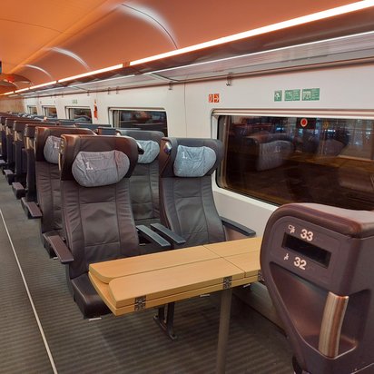 Innenraum eines modernen Fernverkehrszuges in der 1. Klasse. Die Sitze sind mit dunklem Leder bezogen und haben verstellbare Armlehnen sowie Kopfstützen mit hellgrauen Bezügen. In der Mitte befindet sich ein großer Klapptisch mit Steckdosen. Ein digitales Display am Ende des Wagens zeigt die nächste Haltestelle an. Durch das Fenster ist die dunkle Außenumgebung sichtbar.