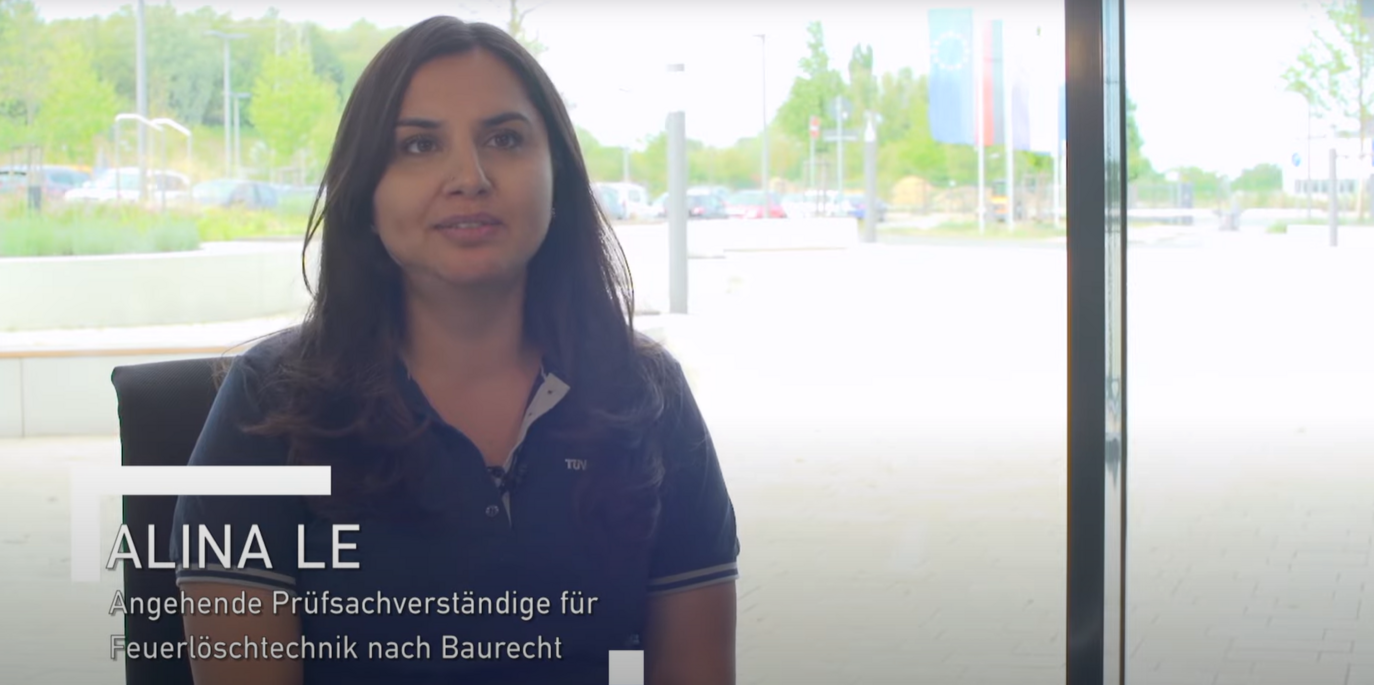 Alina Le, eine Frau mit langen, dunkelbraunen Haaren, sitzt in einem hellen, modernen Raum vor einer großen Fensterfront. Sie trägt ein dunkelblaues Polo-Shirt mit TÜV-Logo und wirkt freundlich und professionell. Ihr Blick ist leicht zur Seite gerichtet, sie spricht in einem Interviewsetting. Im Hintergrund sind geparkte Autos, Grünflächen und mehrere wehende Flaggen zu sehen. Die natürliche Beleuchtung unterstreicht ihre sympathische Ausstrahlung und die offene Atmosphäre des Gesprächs.