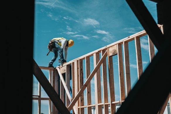 Ein Bauarbeiter mit Sicherheitsweste und gelbem Schutzhelm arbeitet auf einem hölzernen Rohbau eines Gebäudes. Er steht auf einer Balkenkonstruktion und beugt sich nach vorne, während er mit Werkzeug arbeitet. Der Himmel ist blau mit wenigen Wolken im Hintergrund.
