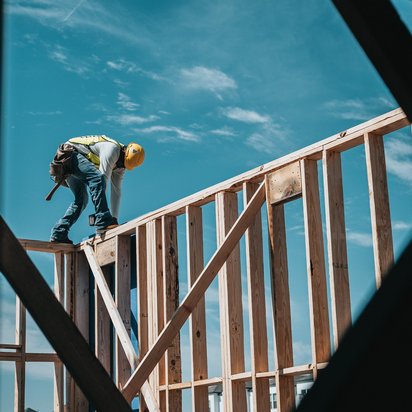 Ein Bauarbeiter mit Sicherheitsweste und gelbem Schutzhelm arbeitet auf einem hölzernen Rohbau eines Gebäudes. Er steht auf einer Balkenkonstruktion und beugt sich nach vorne, während er mit Werkzeug arbeitet. Der Himmel ist blau mit wenigen Wolken im Hintergrund.