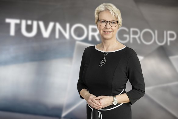 Porträt einer lächelnden Frau mit kurzen, blonden Haaren und Brille. Sie trägt ein elegantes schwarzes Kleid mit weißen Akzenten sowie eine silberne Kette und eine Armbanduhr. Sie steht vor einem unscharfen Hintergrund mit dem Schriftzug „TÜV NORD GROUP“.