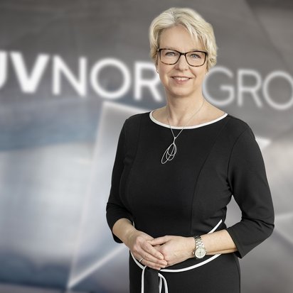 Porträt einer lächelnden Frau mit kurzen, blonden Haaren und Brille. Sie trägt ein elegantes schwarzes Kleid mit weißen Akzenten sowie eine silberne Kette und eine Armbanduhr. Sie steht vor einem unscharfen Hintergrund mit dem Schriftzug „TÜV NORD GROUP“.