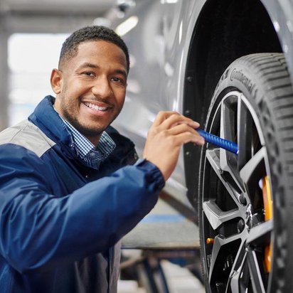 Ein TÜV-Prüfingenieur in blauer Arbeitskleidung überprüft ein Autoreifenprofil mit einer kleinen Taschenlampe. Er lächelt und schaut direkt in die Kamera. Das Fahrzeug steht auf einer Hebebühne in einer Werkstatt mit heller Beleuchtung.