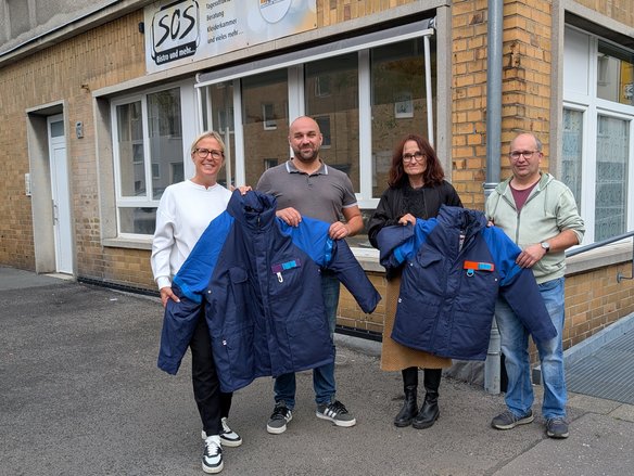 Vier Personen stehen vor dem Gebäude des SOS-Bistros und halten gemeinsam mehrere warme, dunkelblaue Winterjacken mit blauen Ärmeln in die Kamera. Die Gruppe besteht aus zwei Frauen und zwei Männern, die lächeln und sich für die Übergabe der Jacken versammelt haben. Die Frau links trägt eine weiße Jacke und schwarze Hosen, der Mann neben ihr ein graues Poloshirt und Jeans. Die Frau rechts trägt eine schwarze Jacke über einem braunen Kleid mit Stiefeln, während der Mann ganz rechts eine hellgrüne Kapuzenjacke und Jeans trägt. Das Gebäude im Hintergrund hat eine beige Klinkerfassade mit mehreren Fenstern und einem Schild mit der Aufschrift „SOS Bistro und mehr...“.