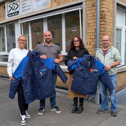 Vier Personen stehen vor dem Gebäude des SOS-Bistros und halten gemeinsam mehrere warme, dunkelblaue Winterjacken mit blauen Ärmeln in die Kamera. Die Gruppe besteht aus zwei Frauen und zwei Männern, die lächeln und sich für die Übergabe der Jacken versammelt haben. Die Frau links trägt eine weiße Jacke und schwarze Hosen, der Mann neben ihr ein graues Poloshirt und Jeans. Die Frau rechts trägt eine schwarze Jacke über einem braunen Kleid mit Stiefeln, während der Mann ganz rechts eine hellgrüne Kapuzenjacke und Jeans trägt. Das Gebäude im Hintergrund hat eine beige Klinkerfassade mit mehreren Fenstern und einem Schild mit der Aufschrift „SOS Bistro und mehr...“.