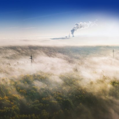 Luftaufnahme einer nebelverhangenen Waldlandschaft mit einem Fabrikschlot, der Rauch in den Himmel abgibt. Der dichte Nebel verdeckt teilweise die Bäume, während Strommasten und -leitungen im Vordergrund sichtbar sind. Über dem Nebel ist der Himmel klar und blau.