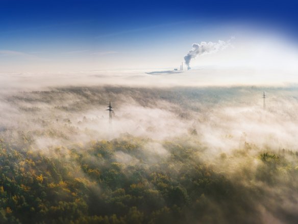 Luftaufnahme einer nebelverhangenen Waldlandschaft mit einem Fabrikschlot, der Rauch in den Himmel abgibt. Der dichte Nebel verdeckt teilweise die Bäume, während Strommasten und -leitungen im Vordergrund sichtbar sind. Über dem Nebel ist der Himmel klar und blau.