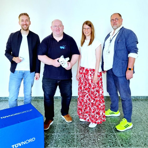 Vier Personen stehen vor einer weißen Wand und lächeln in die Kamera. Sie tragen unterschiedliche Outfits, von leger bis geschäftlich. Der Mann in der Mitte hält ein technisches Gerät in den Händen. Links im Bild steht eine blaue TÜV NORD GROUP-Box. Der Mann ganz rechts trägt ein Namensschild und auffällige neonfarbene Sneakers. Die Szene wirkt entspannt und freundlich.