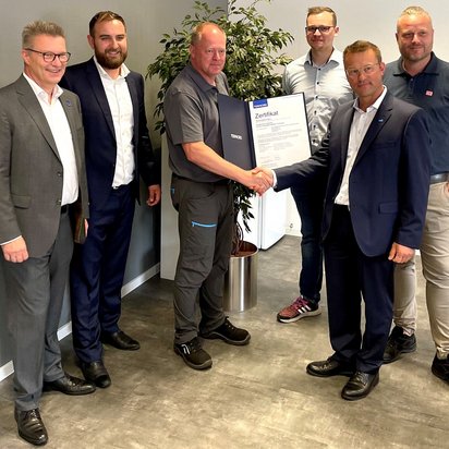 Sechs Männer stehen in einem Büro oder Konferenzraum für ein Zertifikatsübergabe-Foto. Zwei Männer in Anzügen und einer in grauer Arbeitskleidung schütteln sich die Hände, während einer von ihnen ein TÜV NORD-Zertifikat hält. Die anderen Teilnehmer stehen lächelnd daneben. Im Hintergrund befindet sich eine Pflanze und eine helle Wand.