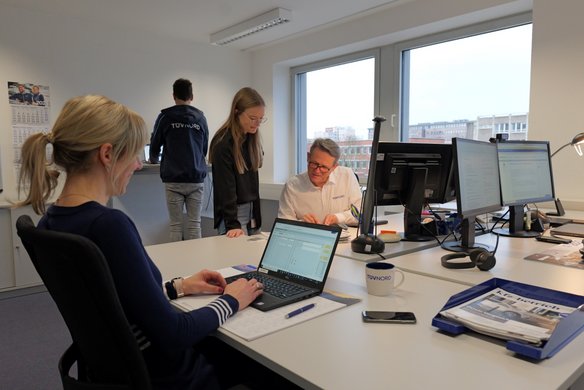 Mehrere Personen arbeiten in einem modernen Büro mit großen Fenstern. Eine Frau tippt auf einem Laptop, während eine Kollegin mit einem älteren Kollegen an Dokumenten arbeitet. Im Hintergrund steht eine Person mit einer TÜV NORD Jacke an einem Stehtisch. Auf den Schreibtischen befinden sich mehrere Bildschirme, Dokumente und eine TÜV NORD-Tasse