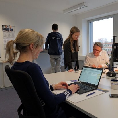 Mehrere Personen arbeiten in einem modernen Büro mit großen Fenstern. Eine Frau tippt auf einem Laptop, während eine Kollegin mit einem älteren Kollegen an Dokumenten arbeitet. Im Hintergrund steht eine Person mit einer TÜV NORD Jacke an einem Stehtisch. Auf den Schreibtischen befinden sich mehrere Bildschirme, Dokumente und eine TÜV NORD-Tasse