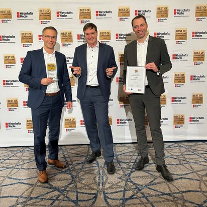 Freuen sich über die Auszeichnung für das Organisationsprojekt NOVA (v.l.n.r.): Jens Neubert (TÜV NORD), Oliver Greiner (Horvath) und Jürgen Terbeck (TÜV NORD). Bild: TÜV NORD Drei Männer in Business-Anzügen stehen auf einer Preisverleihung vor einer Fotowand mit dem Schriftzug „Best of Consulting 2024“ der WirtschaftsWoche. Der Mann links hält eine Trophäe, der Mann rechts eine Urkunde, und der Mann in der Mitte gestikuliert fröhlich. Alle drei lächeln in die Kamera. Der Teppichboden und die professionelle Beleuchtung unterstreichen die feierliche Atmosphäre der Veranstaltung.
