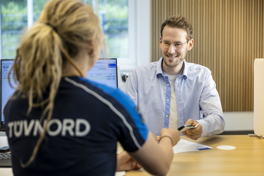 Eine TÜV NORD Mitarbeiterin überreicht einem Kunden Dokumente an einem Schreibtisch.
