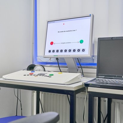 Ein Untersuchungsraum mit einem Reaktionstestgerät für eine medizinisch-psychologische Untersuchung (MPU). Auf einem Schreibtisch steht ein Monitor, der eine Testaufgabe zur Linienverfolgung anzeigt. Vor dem Bildschirm befindet sich ein Steuerpult mit verschiedenen Tasten und Leuchten. Daneben stehen ein Laptop und ein Paar Kopfhörer. Im Vordergrund ist ein blauer Stuhl sichtbar.