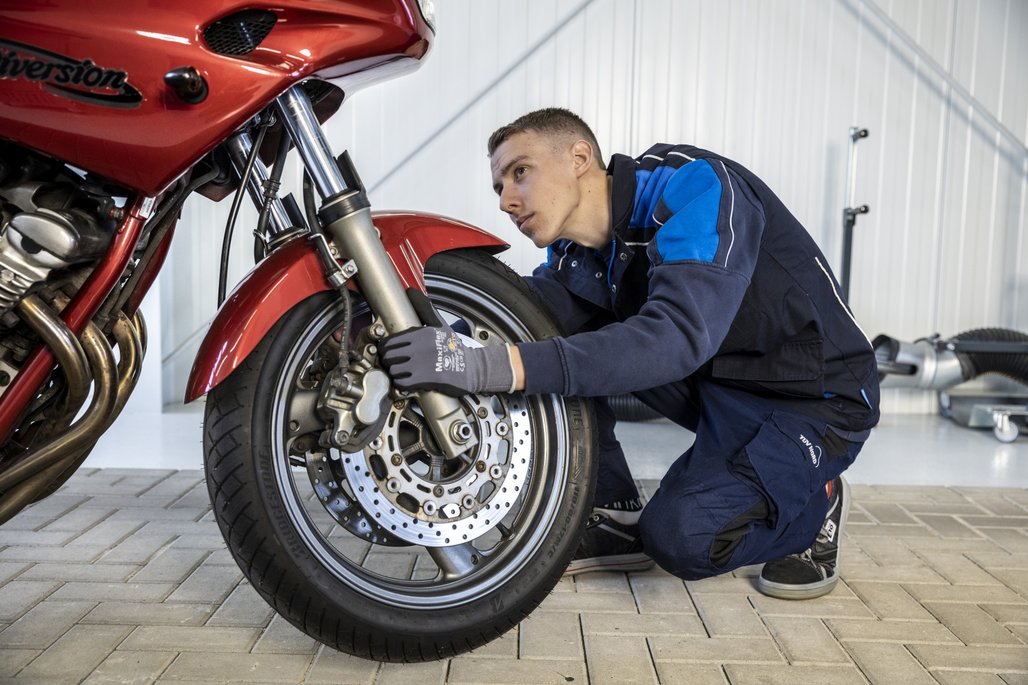 Prüfingenieur von TÜV NORD inspiziert die Bremsen eines roten Motorrads in der Werkstatt, mit Fokus auf Bremsscheiben und Vorderradaufhängung.