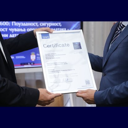 Zwei Männer in Anzügen halten gemeinsam ein gerahmtes TÜV-Zertifikat. Das Zertifikat bestätigt die Sicherheitsstandards eines IT- und eGovernment-Büros in Serbien nach EN 50600. Im Hintergrund ist ein Bildschirm mit serbischer Beschriftung und einer Landesflagge zu sehen.