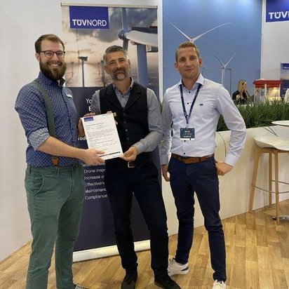 Drei Männer stehen an einem TÜV NORD-Messestand auf einer Fachmesse für Windenergie. Sie halten gemeinsam ein Zertifikat und lächeln in die Kamera. Der Mann links trägt ein blaues Hemd mit grünen Hosenträgern und eine grüne Hose. Der Mann in der Mitte hat graue Haare und einen Bart, trägt eine dunkle Weste über einem Hemd. Der Mann rechts trägt ein weißes Hemd mit Namensschild und eine dunkle Hose. Im Hintergrund sind Banner mit Windkraftanlagen sowie Messebesucher und Sitzgelegenheiten zu sehen.