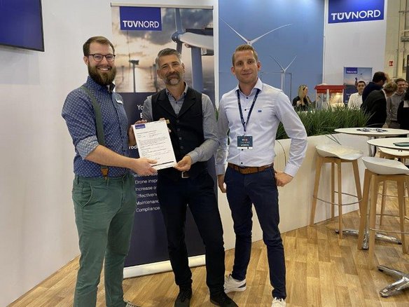 Drei Männer stehen an einem TÜV NORD-Messestand auf einer Fachmesse für Windenergie. Sie halten gemeinsam ein Zertifikat und lächeln in die Kamera. Der Mann links trägt ein blaues Hemd mit grünen Hosenträgern und eine grüne Hose. Der Mann in der Mitte hat graue Haare und einen Bart, trägt eine dunkle Weste über einem Hemd. Der Mann rechts trägt ein weißes Hemd mit Namensschild und eine dunkle Hose. Im Hintergrund sind Banner mit Windkraftanlagen sowie Messebesucher und Sitzgelegenheiten zu sehen.