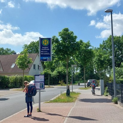 Eine Bushaltestelle in einer Wohngegend mit dem Schild „Burgdorf Havelstraße“ für die Buslinie 905. Eine Schülerin mit einem großen Rucksack steht an der Haltestelle und schaut auf den Fahrplan. Im Hintergrund fährt ein weiteres Kind mit einem Fahrrad auf dem Gehweg. Die Umgebung ist geprägt von Einfamilienhäusern mit roten Ziegeldächern, grünen Bäumen und einer ruhigen Straße unter blauem Himmel mit einigen Wolken.
