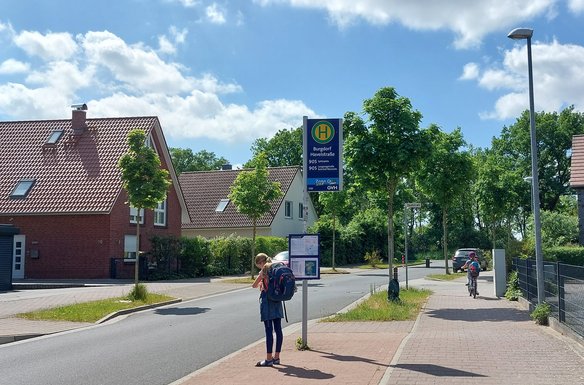 Eine Bushaltestelle in einer Wohngegend mit dem Schild „Burgdorf Havelstraße“ für die Buslinie 905. Eine Schülerin mit einem großen Rucksack steht an der Haltestelle und schaut auf den Fahrplan. Im Hintergrund fährt ein weiteres Kind mit einem Fahrrad auf dem Gehweg. Die Umgebung ist geprägt von Einfamilienhäusern mit roten Ziegeldächern, grünen Bäumen und einer ruhigen Straße unter blauem Himmel mit einigen Wolken.