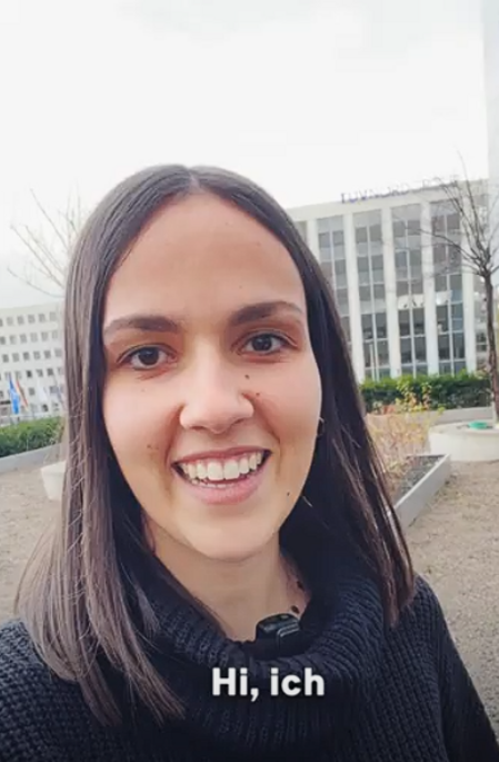Das Bild zeigt Ricarda, eine Recruiterin, die lächelnd in die Kamera blickt. Sie hat schulterlanges dunkelbraunes Haar und trägt einen schwarzen Pullover. Im Hintergrund ist ein modernes Bürogebäude mit dem Schriftzug ‚TÜV NORD‘ sichtbar. Die Szene spielt sich im Freien ab, auf einer Fläche mit bepflanzten Hochbeeten, kahlen Bäumen und Gehwegen. Das Bild hat einen leichten Selfie-Charakter, und im unteren Bereich ist der Schriftzug ‚Hi, ich‘ eingeblendet, was darauf hindeutet, dass es sich um den Beginn eines Testimonial-Videos handelt. Dieses Thumbnail gehört zu einem Mitarbeiter-Testimonial und vermittelt eine offene, einladende und professionelle Atmosphäre im Bereich Recruiting.
