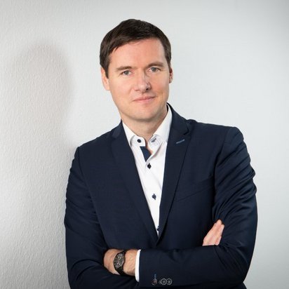 Ein Mann mit kurzen, dunklen Haaren steht mit verschränkten Armen vor einer weißen Wand. Er trägt einen dunkelblauen Anzug mit einem weißen Hemd, das dunkle Knöpfe und ein dezentes Muster aufweist. Sein Blick ist freundlich und selbstbewusst.