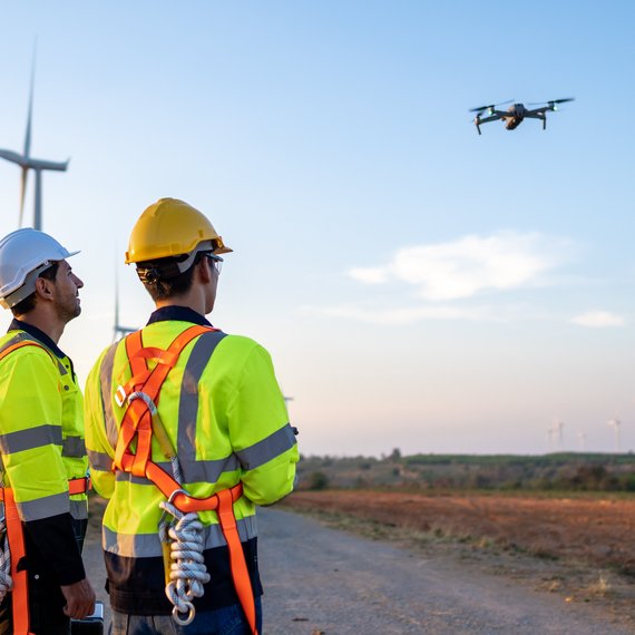 Two engineers in safety gear operate a drone near a wind farm during sunset.