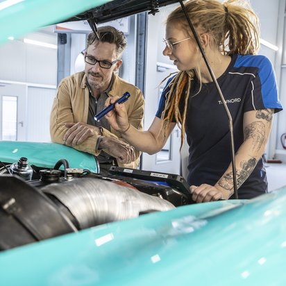 Mann und Frau im TÜV NORD T-Shirt begutachten ein Auto mit geöffneter Motorhaube.