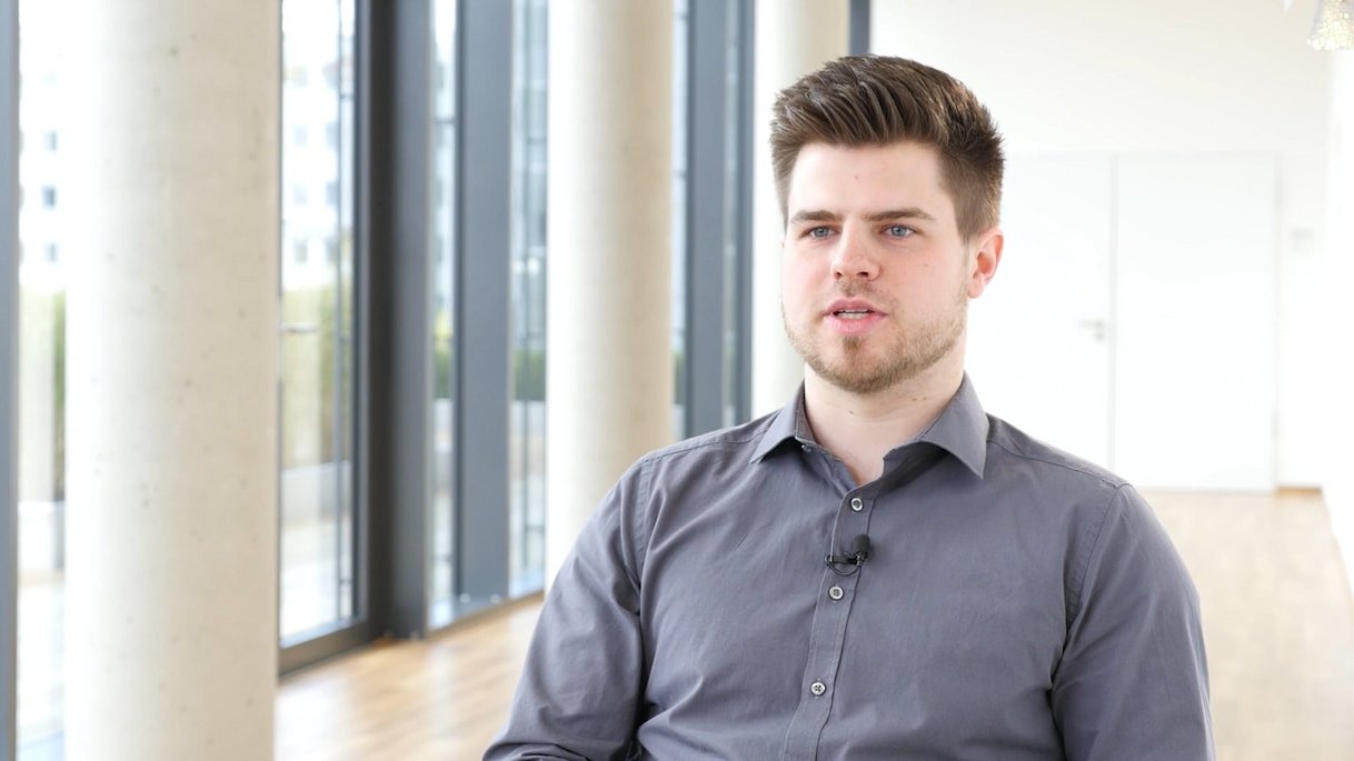 Das Bild zeigt Philipp Tetzlaff, einen Mitarbeiter im Bereich interne IT-Dienstleistungen, in einem modernen, lichtdurchfluteten Büro. Er trägt ein graues Hemd und ein dezentes Ansteckmikrofon. Sein Gesichtsausdruck ist aufmerksam und konzentriert, während er leicht an der Kamera vorbeischaut. Der Hintergrund mit großen Fenstern und hellen Säulen sorgt für eine offene und professionelle Atmosphäre. Dieses Thumbnail gehört zu einem Mitarbeiter-Testimonial und hebt die Rolle von Max Mustermann in der internen IT des Unternehmens hervor.