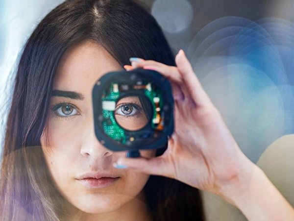 A woman holds a computer board in front of her left eye and looks through a hole in it into the camera
