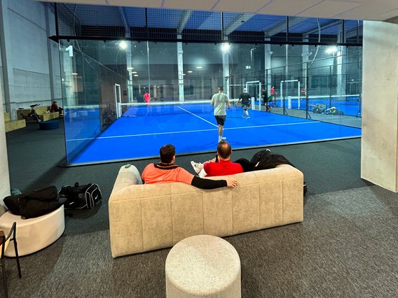 Zwei Personen sitzen auf einem grauen Sofa in einer modernen Padelhalle und schauen durch eine Glaswand auf ein laufendes Spiel auf einem blauen Padelplatz. Im Hintergrund sind weitere Spieler auf benachbarten Plätzen aktiv, während die Halle mit Betonwänden und künstlicher Beleuchtung eine industrielle Atmosphäre vermittelt.