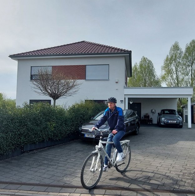 Ein Mann mit Fahrradhelm und blauer Jacke fährt mit einem weißen Fahrrad aus der Einfahrt eines modernen Einfamilienhauses. Im Hintergrund sind eine Hecke, mehrere Autos und eine Ladestation für ein Elektrofahrzeug zu sehen