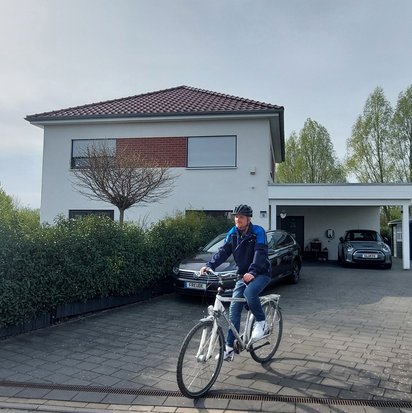 Ein Mann mit Fahrradhelm und blauer Jacke fährt mit einem weißen Fahrrad aus der Einfahrt eines modernen Einfamilienhauses. Im Hintergrund sind eine Hecke, mehrere Autos und eine Ladestation für ein Elektrofahrzeug zu sehen.