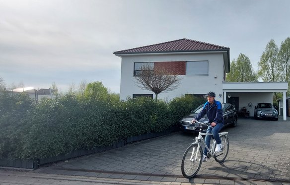 Ein Mann mit Fahrradhelm und blauer Jacke fährt mit einem weißen Fahrrad aus der Einfahrt eines modernen Einfamilienhauses. Im Hintergrund sind eine Hecke, mehrere Autos und eine Ladestation für ein Elektrofahrzeug zu sehen