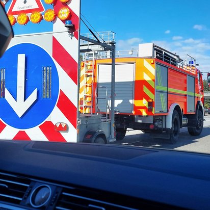 Ein Blick aus dem Inneren eines Autos auf eine Autobahn. Im Vordergrund das Lenkrad und das Armaturenbrett, im Mittelgrund ein großer, rot-weiß gestreifter Verkehrsschild mit einem Pfeil nach unten, der auf eine Baustelle hinweist. Dahinter ein orangefarbener LKW der Feuerwehr mit Blaulicht, gefolgt von anderen Fahrzeugen. Am rechten Bildrand ein Motorradfahrer und ein grüner Kastenwagen
