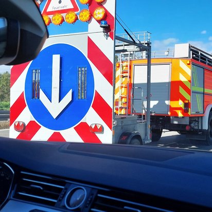 Ein Blick aus dem Inneren eines Autos auf eine Autobahn. Im Vordergrund das Lenkrad und das Armaturenbrett, im Mittelgrund ein großer, rot-weiß gestreifter Verkehrsschild mit einem Pfeil nach unten, der auf eine Baustelle hinweist. Dahinter ein orangefarbener LKW der Feuerwehr mit Blaulicht, gefolgt von anderen Fahrzeugen. Am rechten Bildrand ein Motorradfahrer und ein grüner Kastenwagen