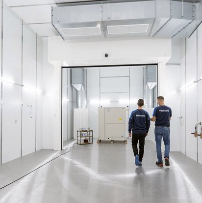 Zwei Mitarbeitende von TÜV NORD gehen durch einen großen, hell beleuchteten Prüfraum mit weißen Wänden und modernen Lüftungssystemen. Sie tragen dunkelblaue Arbeitskleidung mit dem TÜV NORD-Logo auf dem Rücken. Im Hintergrund befindet sich eine große technische Anlage, vermutlich eine Wärmepumpe, die für Tests vorbereitet wird. Der Raum wirkt industriell und ist mit technischen Geräten ausgestattet.