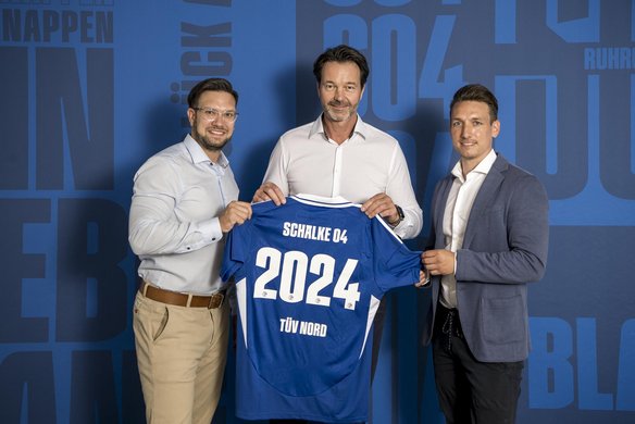 Drei Männer in Business-Kleidung stehen vor einer blauen Wand mit Schriftzügen und dem Logo von Schalke 04. Sie halten gemeinsam ein blaues Fußballtrikot mit der Aufschrift „Schalke 04“, der Jahreszahl „2024“ und dem TÜV NORD-Logo. Die Männer lächeln in die Kamera, während sie das Trikot präsentieren.