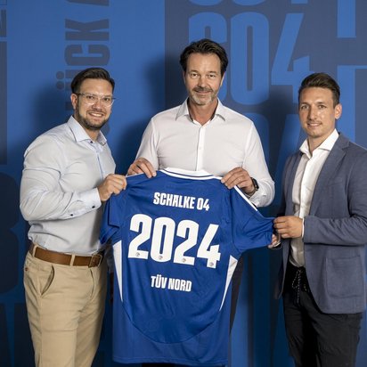 Drei Männer in Business-Kleidung stehen vor einer blauen Wand mit Schriftzügen und dem Logo von Schalke 04. Sie halten gemeinsam ein blaues Fußballtrikot mit der Aufschrift „Schalke 04“, der Jahreszahl „2024“ und dem TÜV NORD-Logo. Die Männer lächeln in die Kamera, während sie das Trikot präsentieren.